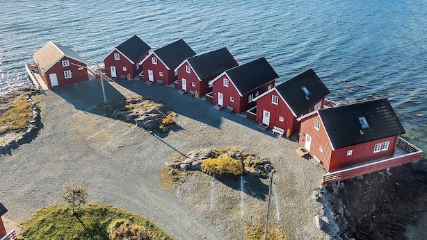 Une rangée de maisons en bois peintes en rouge, avec des toits noirs, situées sur une côte rocheuse au bord de la mer.