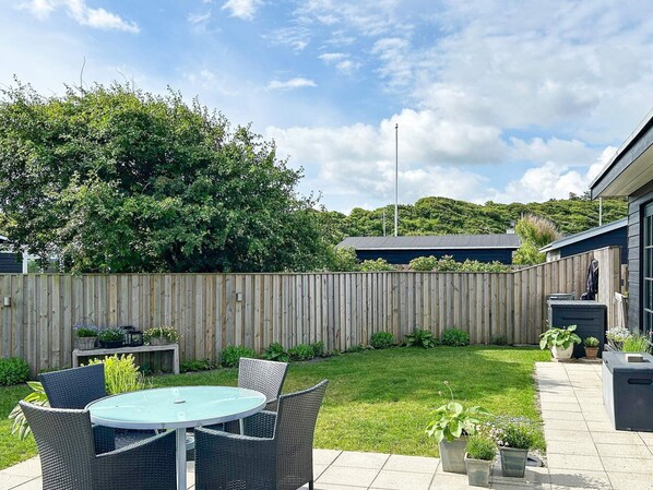 A backyard with a round table and wicker chairs, surrounded by a wooden fence and lush greenery.