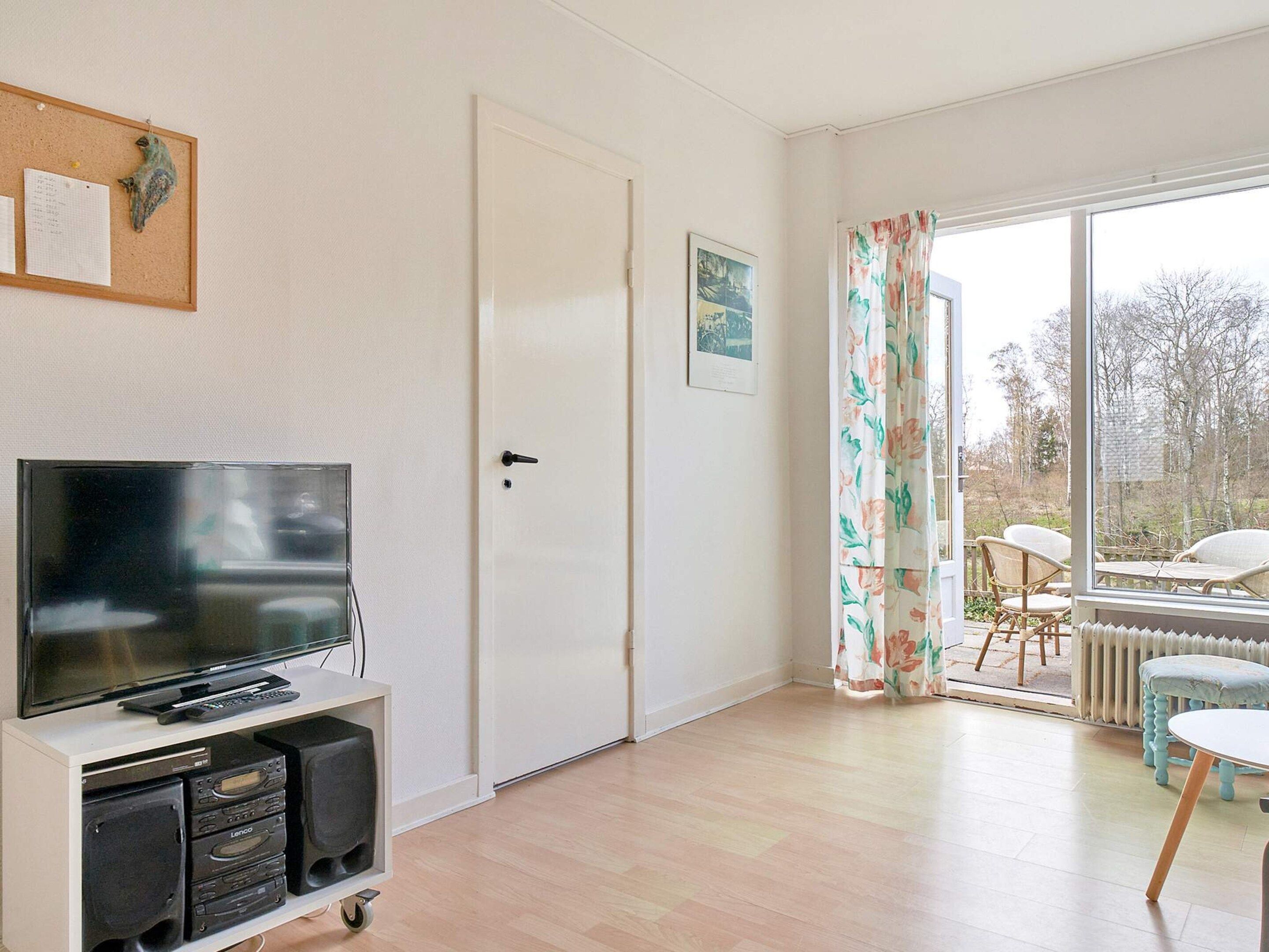 A room with a television on a stand, a door, a window with curtains, and a view of the outside.