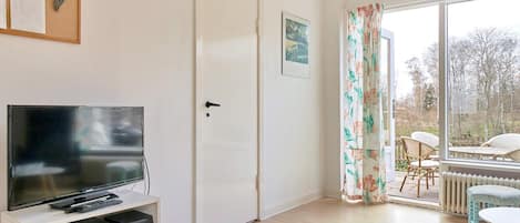 A room with a television on a stand, a door, a window with curtains, and a view of the outside.
