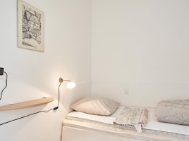 Quarto com uma cama de solteiro, luminária de cabeceira, prateleira na parede e um quadro emoldurado.