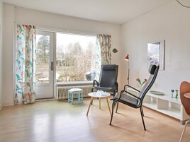 Una habitación con un ventanal grande, un radiador, una silla negra, un taburete pequeño y una mesa blanca.