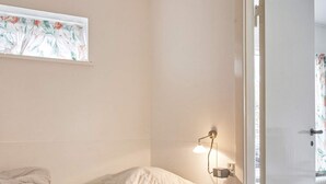 A bedroom with a bed, a nightstand, a wall-mounted lamp, and a window with a floral curtain. - 2 Person Holiday Home in Aakirkeby (Akirkeby)