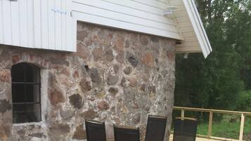 Pared exterior de piedra con una ventana y una terraza de madera con sillas.