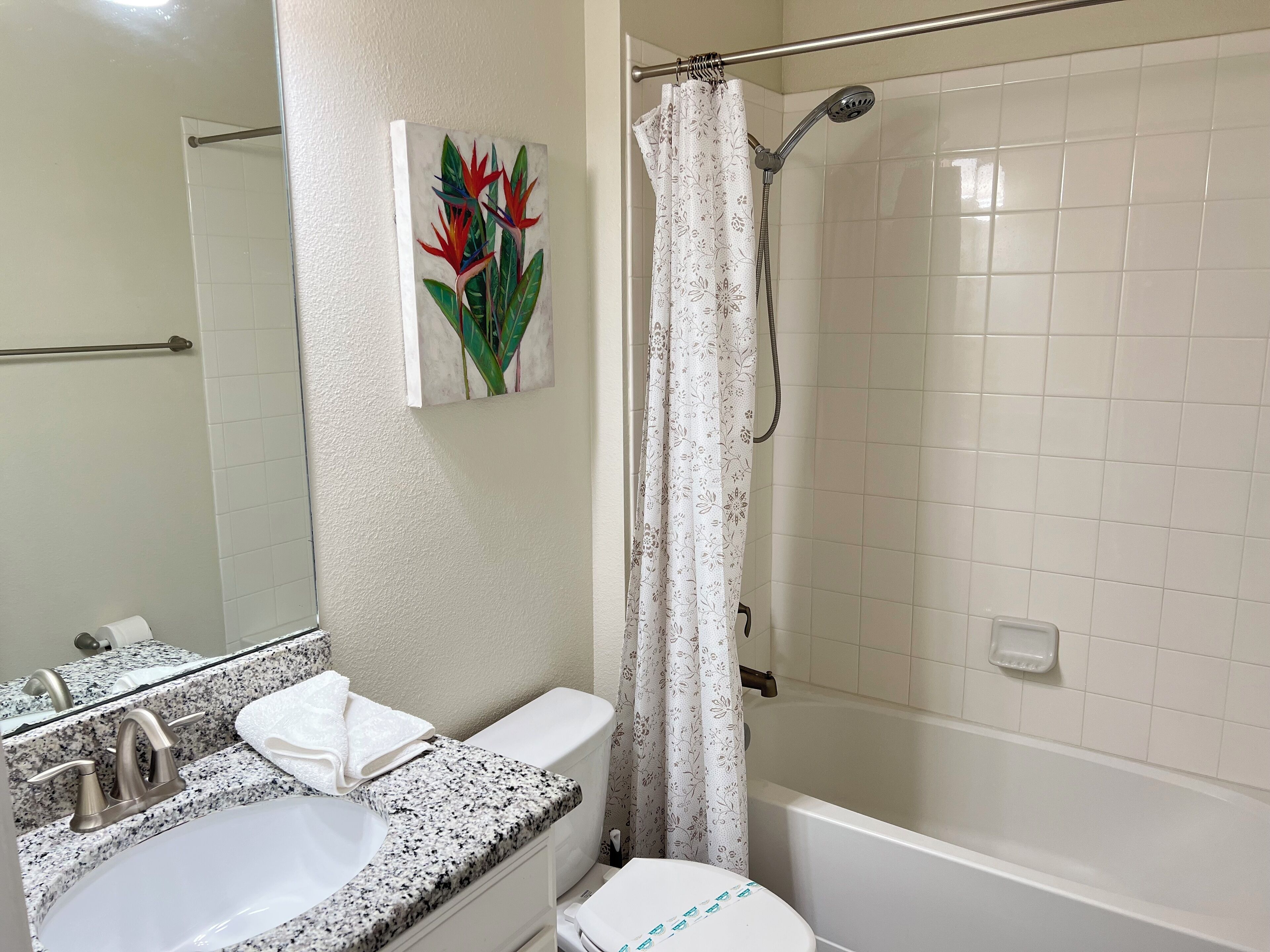 Une salle de bain avec un lavabo sur un comptoir en granit, des toilettes, une douche avec un rideau de douche à motifs floraux et un tableau représentant des fleurs tropicales au mur.