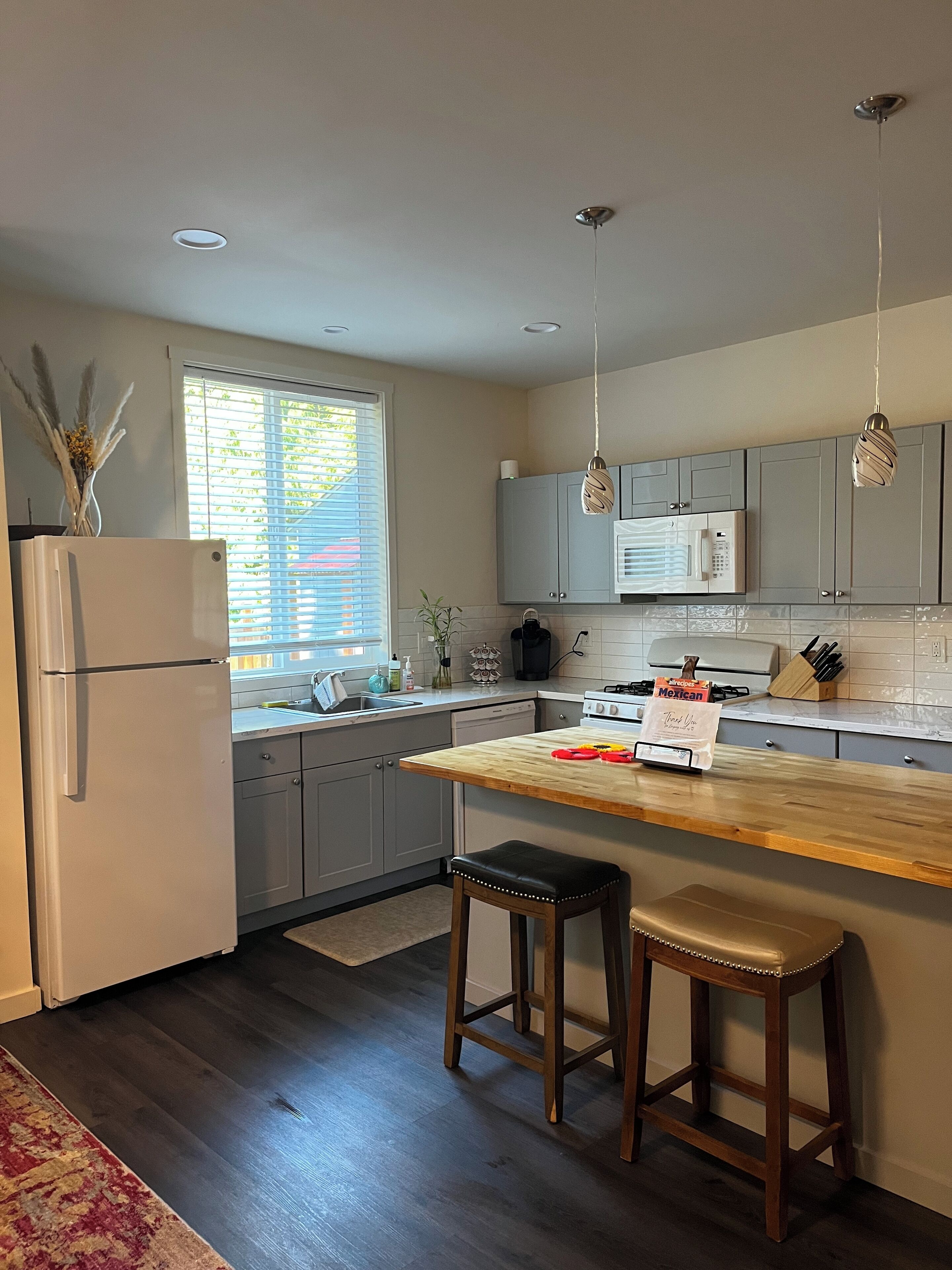 A modern kitchen with grey cabinets, a wooden countertop, two stools, a refrigerator, a window with blinds, and pendant lights.