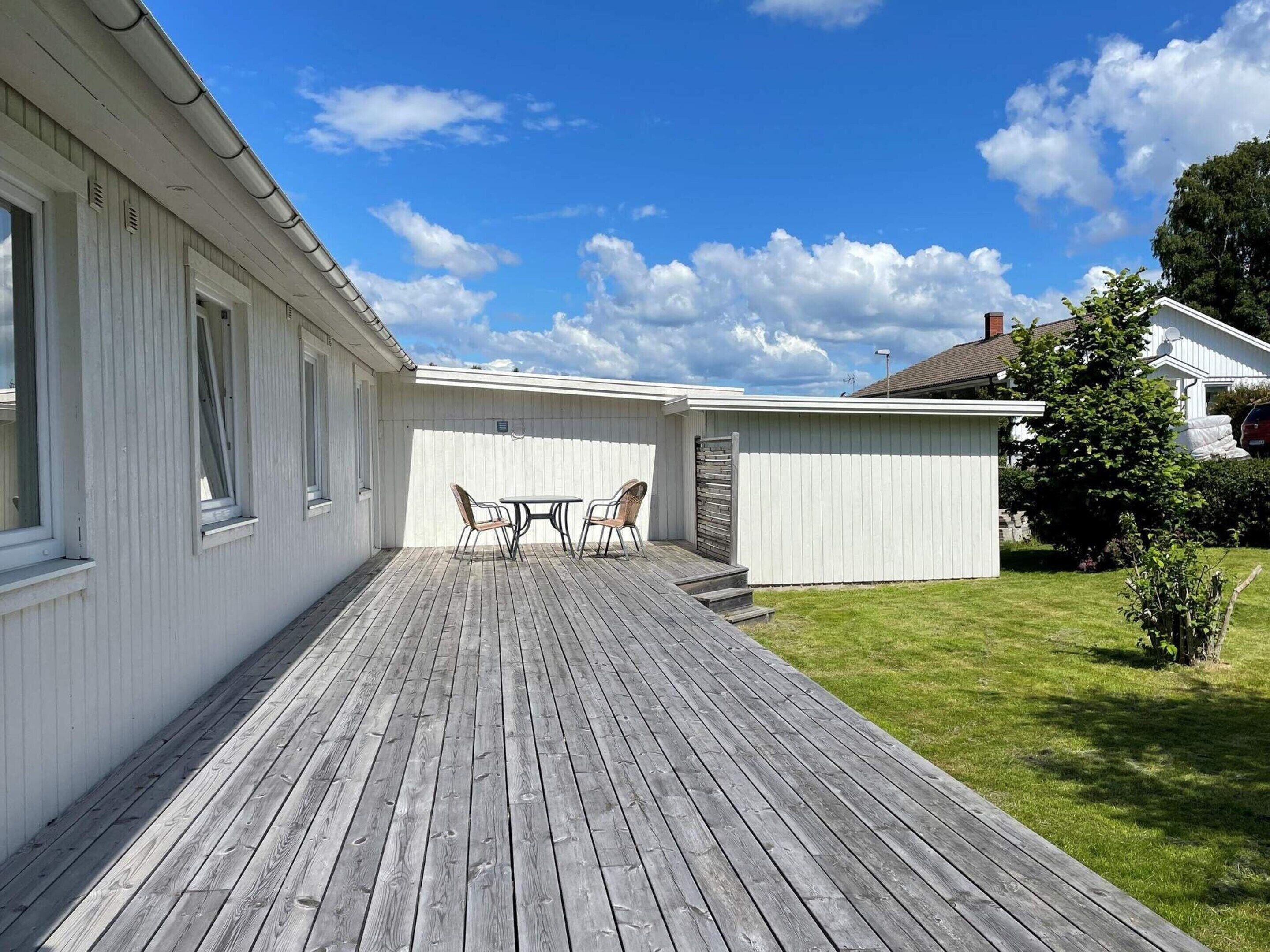 A wooden deck with a table and chairs, a white building with windows, and a grassy yard.