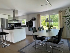 A modern kitchen with a dining area, large windows, and a view of greenery.