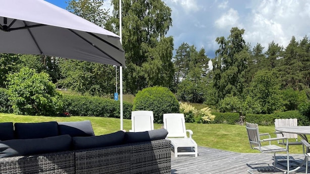 Een terras buiten met een rietenbank, witte stoelen en een tafel.