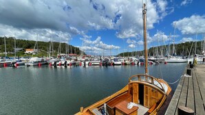 Een houten boot afgemeerd in een jachthaven, met diverse andere boten, een houten steiger en op de achtergrond huizen en bomen onder een lichtbewolkte lucht.