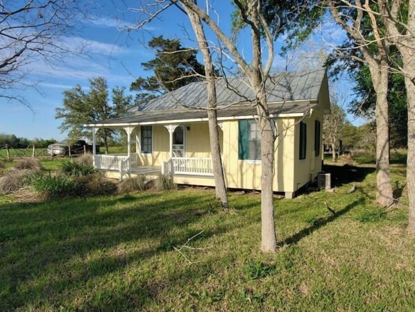 A small, single-story house with a porch, surrounded by trees and grass.