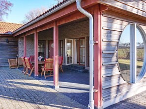A covered patio with wooden chairs and a table, a brick wall, and a large window. - 4 Star Holiday Home in Morbylanga (Morbylanga)
