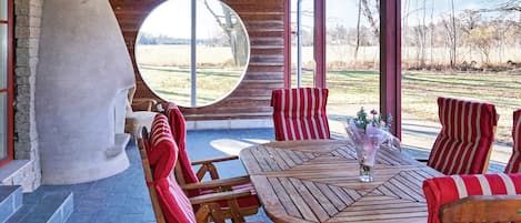 A covered outdoor seating area with a round table and chairs, a large window, and a stone structure.