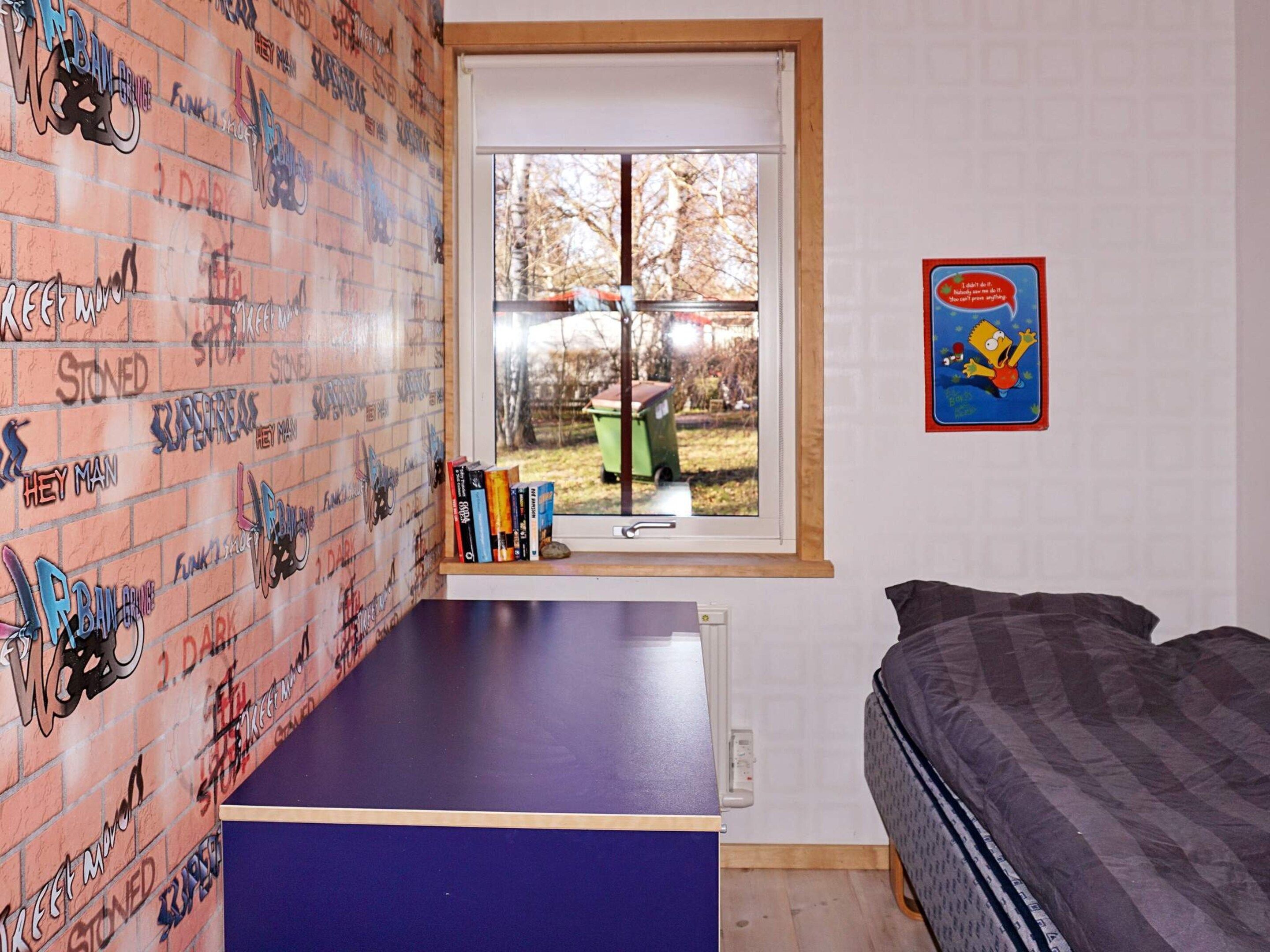 Een kamer met een wand met bakstenen patroon, een raam met uitzicht op een binnentuin, een bed en een klein tafeltje met boeken.