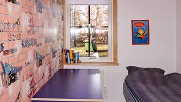 A room with a brick-patterned wall, a window with a view of a yard, a bed, and a small table with books.