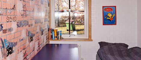 A room with a brick-patterned wall, a window with a view of a yard, a bed, and a small table with books.