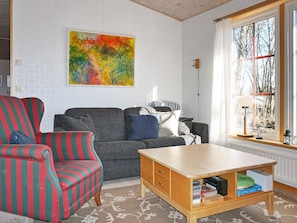 A living room with a sofa, armchair, and coffee table. There is a painting on the wall and a window with a view of trees.