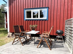 A wooden deck with outdoor furniture, a barbecue grill, and a window with a blue frame. - 4 Person Holiday Home in Gamleby (Gamleby)