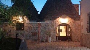 A stone building with a conical roof and a lit entrance, surrounded by a paved courtyard and greenery. - TRULLI ALLEGRETTI (Private pool): your vacation in the heart of Puglia (Noci)