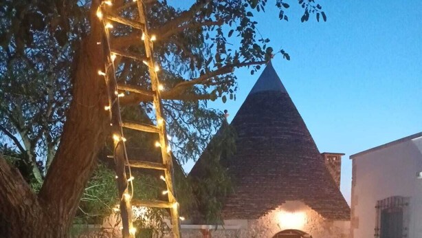 A stone building with a conical roof, a wooden ladder with string lights, and a paved courtyard. - TRULLI ALLEGRETTI (Private pool): your vacation in the heart of Puglia (Noci)