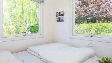 A bedroom with a bed, two windows, a small table with a plant, and a wall-mounted power outlet.