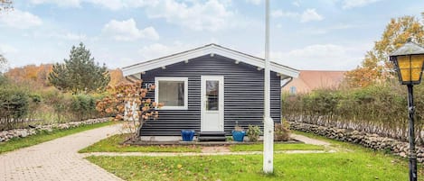 Une petite cabane aux couleurs sombres, dotée d’une porte et d’une fenêtre blanches, entourée d’un mur de pierres et bordée d’un chemin pavé.