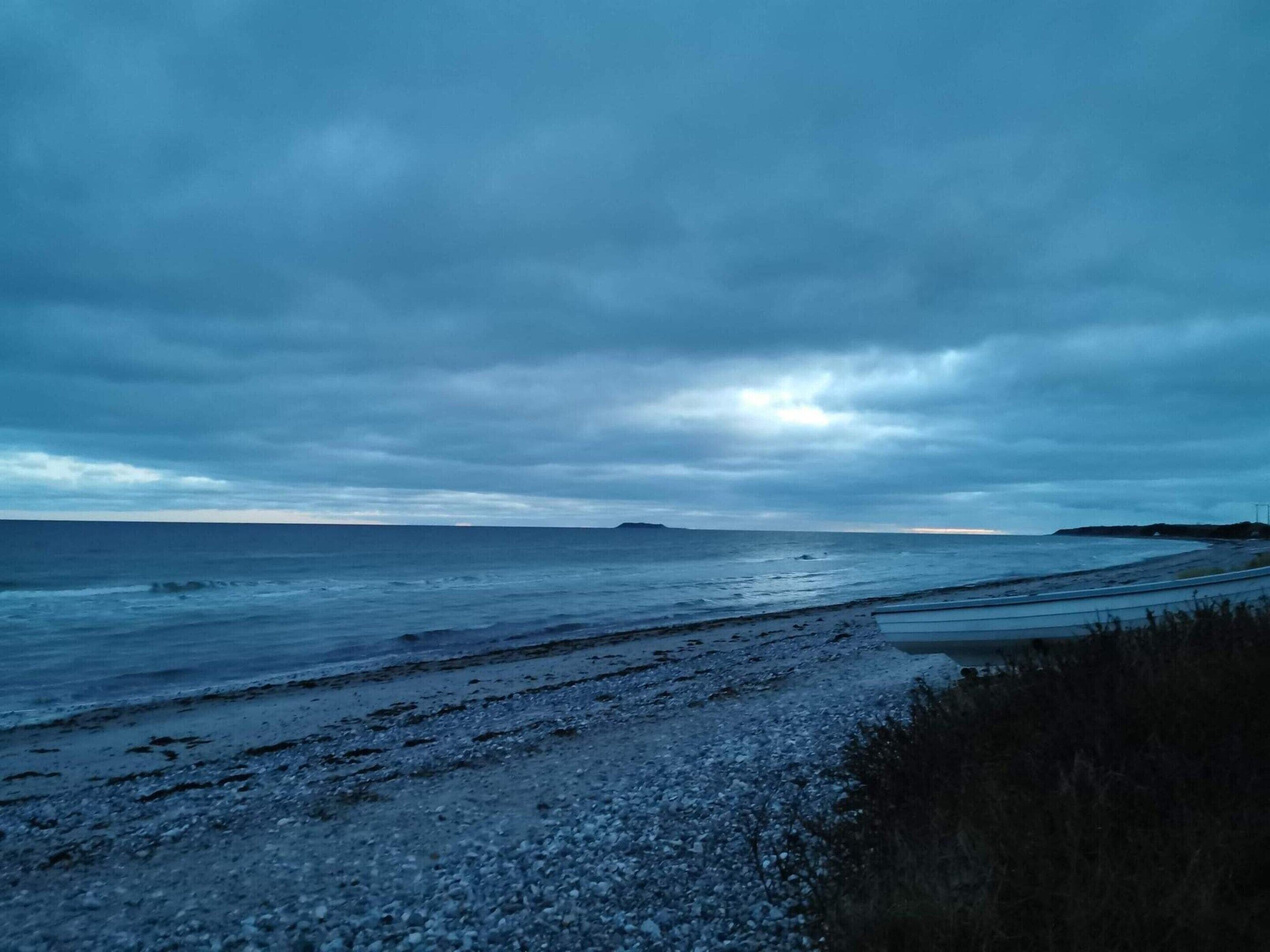 Une route côtière bordée d’une plage de galets, avec une vue lointaine sur la mer sous un ciel nuageux.
