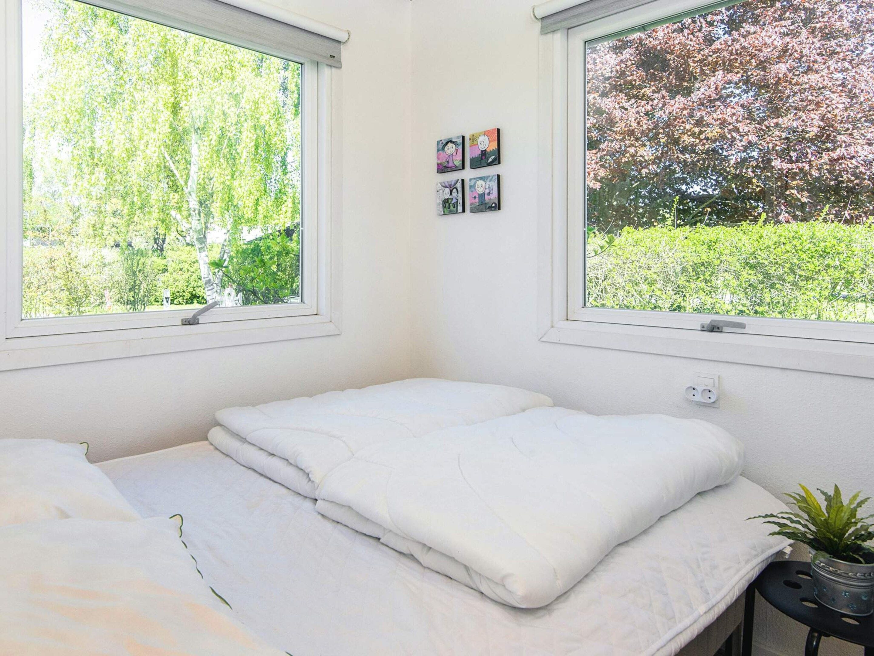 Un dormitorio con cama, dos ventanas, un enchufe en la pared y una pequeña planta en maceta.