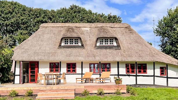 Una casa tradicional con techo de paja, fachada blanca y detalles rojos, que cuenta con un patio con mobiliario exterior y un pequeño jardín.