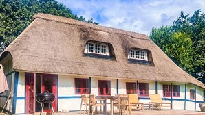 Una casa con techo de paja, terraza de madera, ventanas de marco rojo y una parrilla.