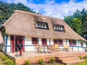 Une maison à toit de chaume, dotée d’une terrasse en bois, de fenêtres à cadre rouge et d’un barbecue.