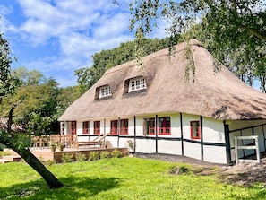 Une maison traditionnelle à toit de chaume, avec une façade blanche et des encadrements de fenêtres rouges, entourée de verdure.