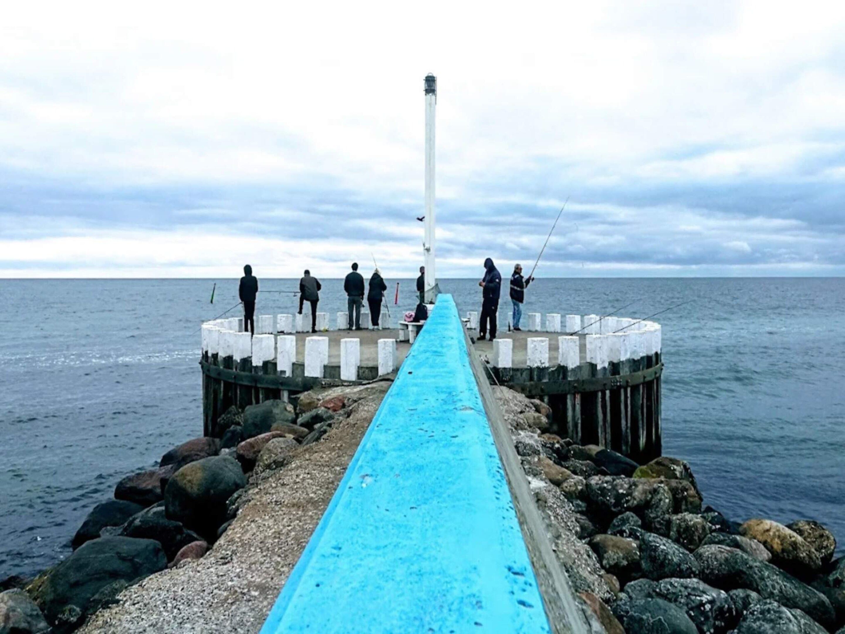 Un muelle se adentra en el agua, con un paseo marítimo azul, rodeado de rocas y un faro al fondo.