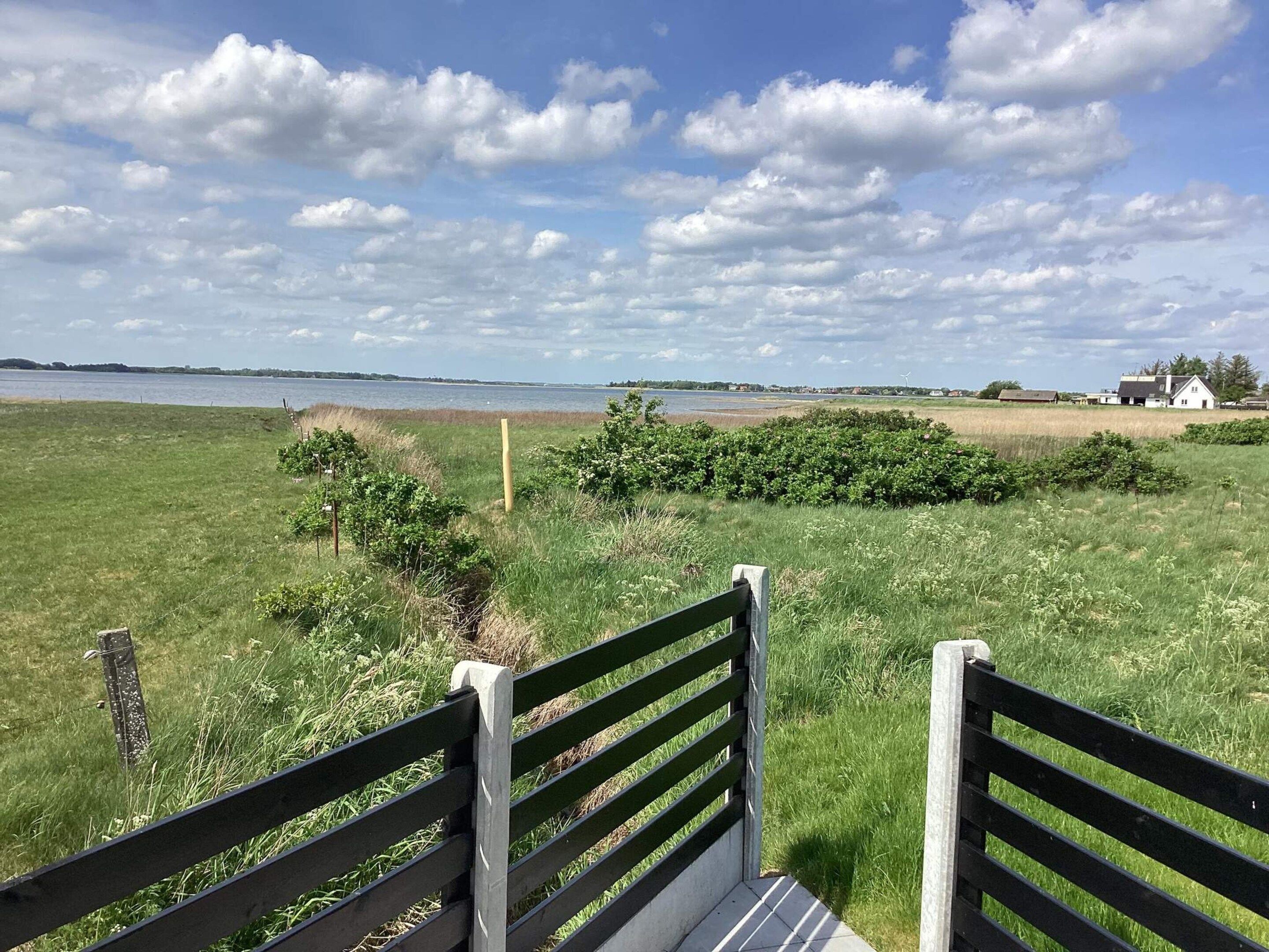 Ein grünes Feld mit einer Bank im Vordergrund und einem Gewässer im Hintergrund.