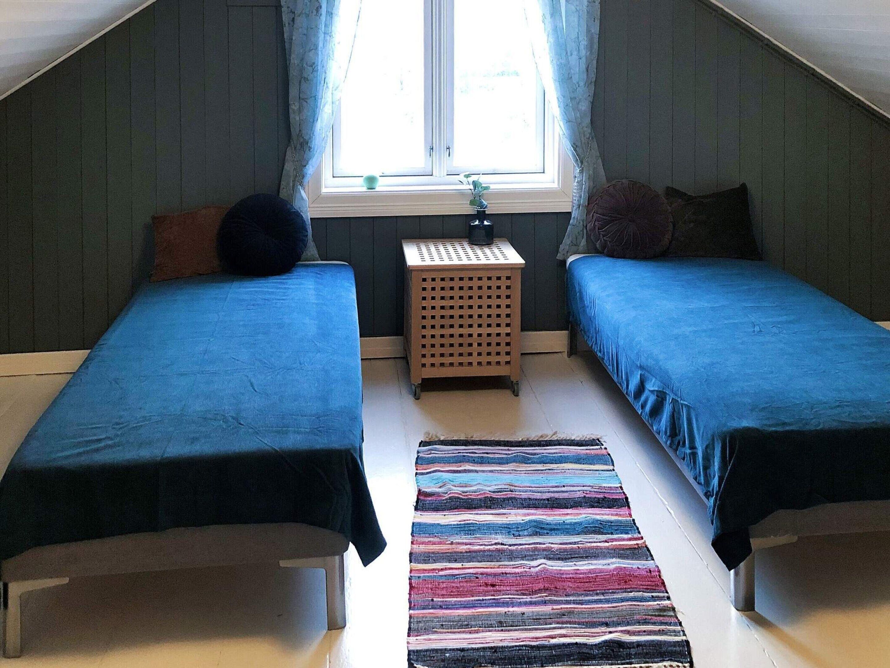 A room with two single beds, a small wooden side table, a colorful striped rug, and a window with curtains.