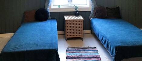 A room with two single beds, a small wooden side table, a colorful striped rug, and a window with curtains.
