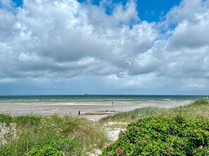En strand med klart skys og en person, der flyver en drage, samt et skib i det fjerne.