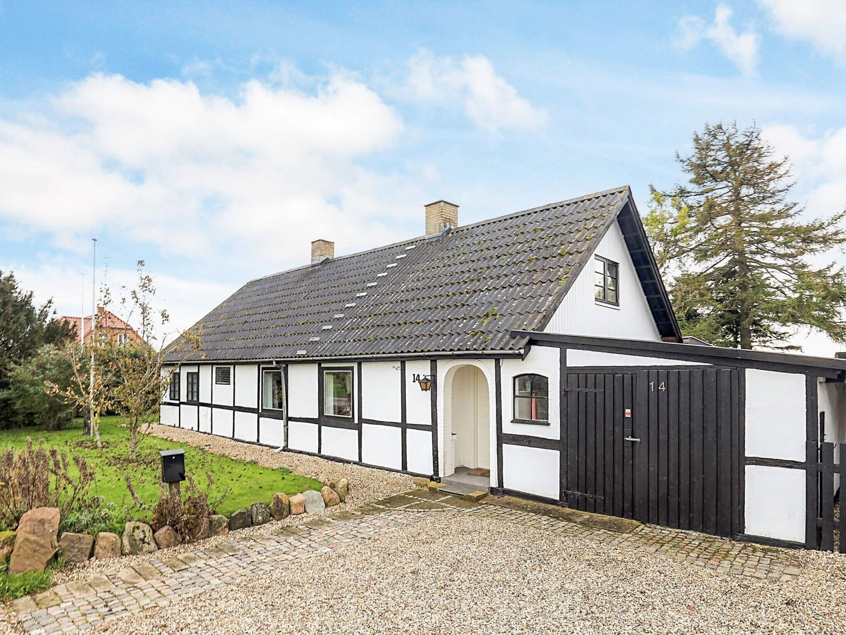 A traditional half-timbered house with a steeply pitched roof, a black door, and a gravel driveway leading to a white entrance.