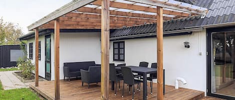 A covered outdoor patio with a wooden pergola, a dining table with chairs, and a white exterior wall.
