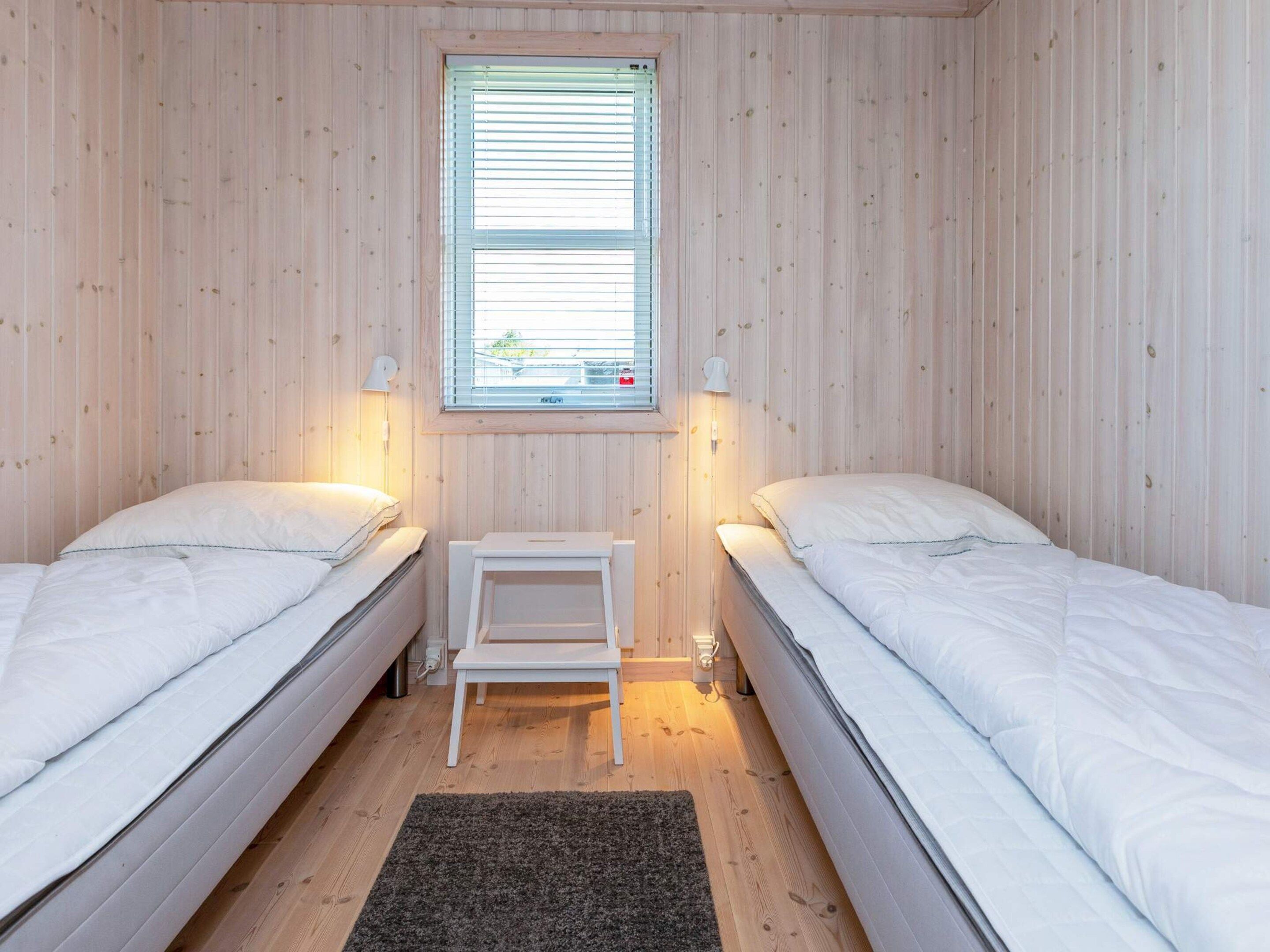 A room with two single beds, a small white nightstand, a window with blinds, and a wall-mounted lamp.