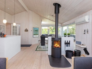 A modern interior with a central black wood-burning stove, wooden walls, a kitchen area, and a seating space with a view of greenery outside. - 7 Person Holiday Home in Logstor (Logstor)