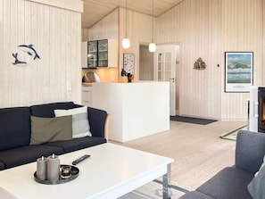 A modern living room with a white coffee table, a black sofa, and a gray armchair. There is a kitchen area with a white cabinet and a hanging light fixture.