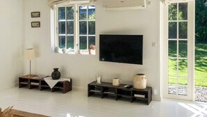 A spacious living room with a large flat-screen TV mounted on the wall, a wooden coffee table, and a large window providing a view of a green garden. - 6 Person Holiday Home in Gilleleje (Gilleleje)
