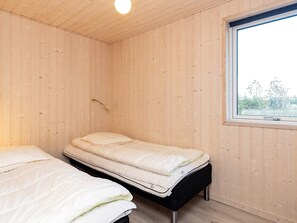 Une pièce aux murs en bois, avec un lit simple doté d’un matelas, une table de chevet et une fenêtre donnant sur un paysage verdoyant.
