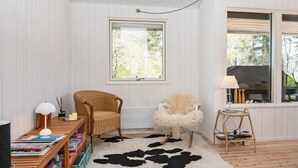 A cozy room with a wooden floor, a cowhide rug, a wicker chair, a fluffy chair, a small table with a lamp, and a bookshelf. - 6 Person Holiday Home in Ebeltoft (Ebeltoft)
