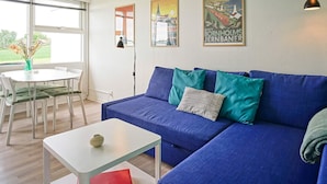 Una sala de estar moderna con un sofá azul, una mesa de centro blanca y un comedor con una mesa y sillas blancas. Hay dos cuadros en la pared y una ventana con persianas.