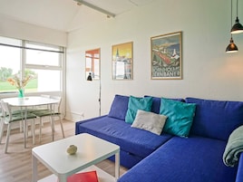 Una sala de estar moderna con un sofá azul, una mesa de centro blanca y un comedor con una mesa y sillas blancas. Hay dos cuadros en la pared y una ventana con persianas.
