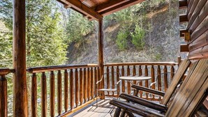 A wooden deck with a table and chairs, overlooking a forested hillside.