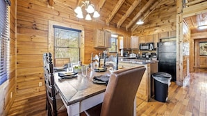 A wooden cabin interior with a dining table set for four, a kitchen area with appliances, and a window with blinds.
