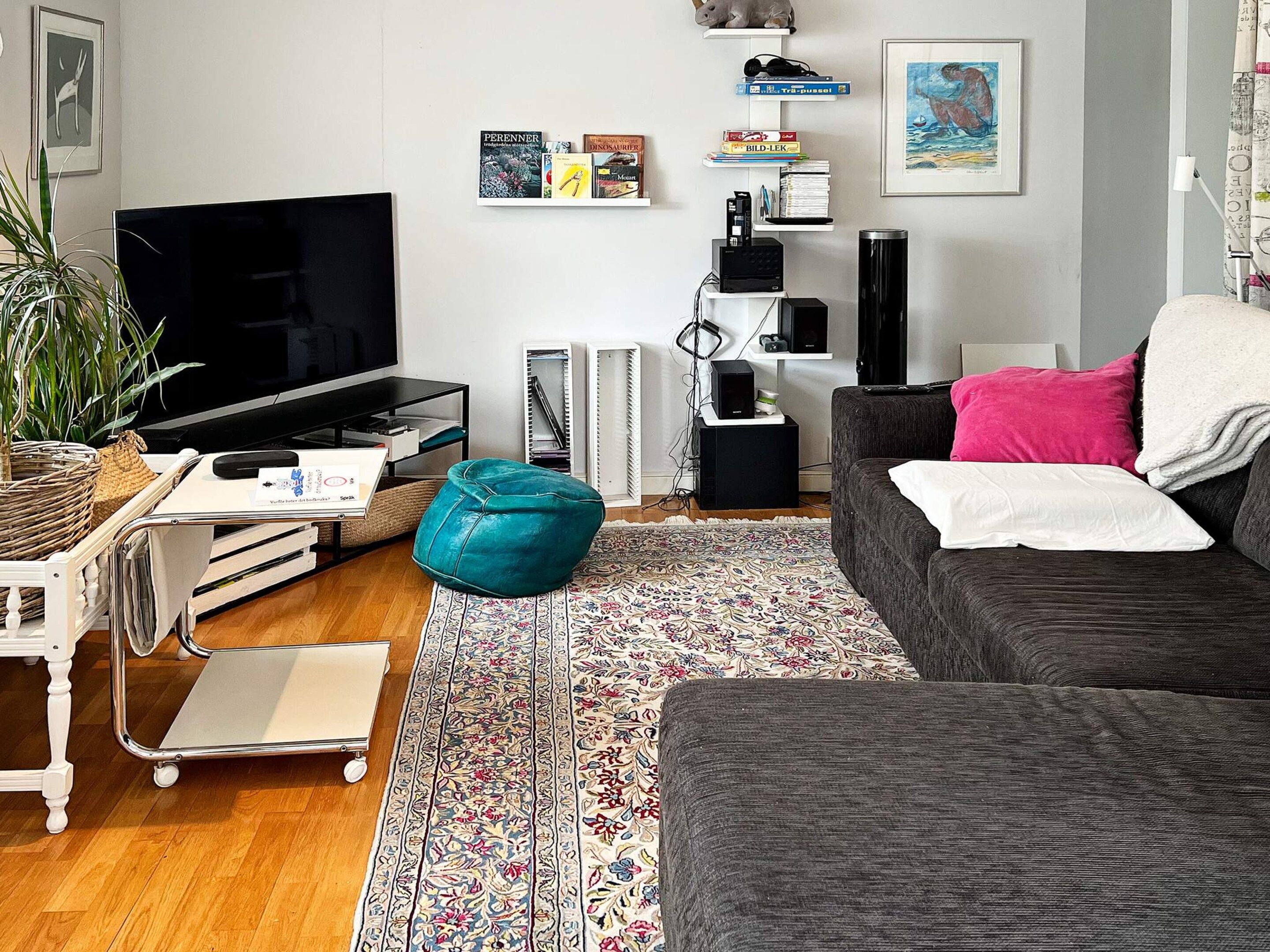A living room with a flat-screen TV, a sofa, a teal ottoman, a patterned rug, and a bookshelf with books and decorative items.